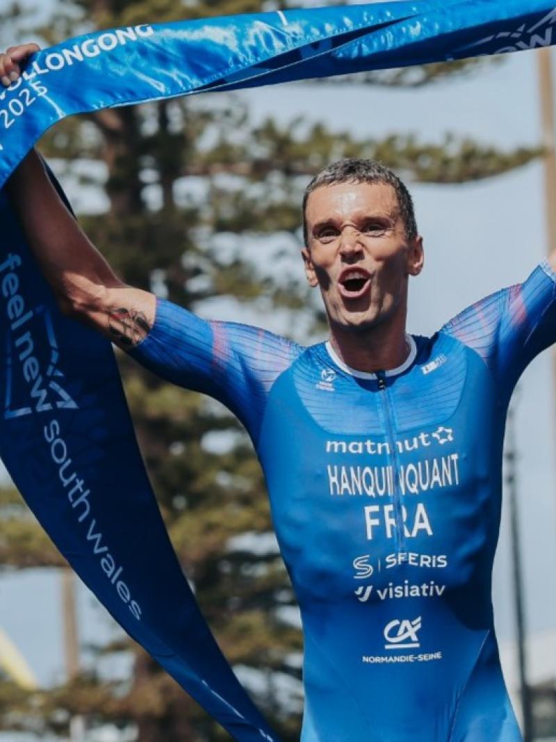 A male Para triathlon athlete wearing a blue track suit holds a blue finish tape and celebrates
