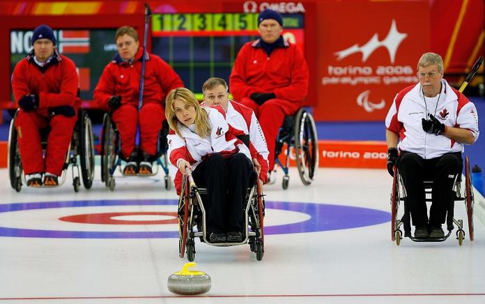 A female wheelchair curling athlete delivers a yellow curling stone using a long stick, while her teammate, a female player, is holding her wheelchair from behind