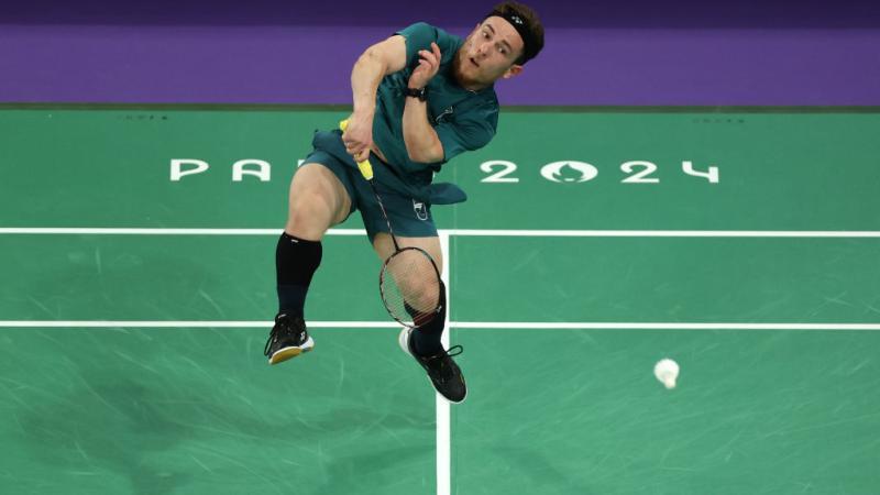 A male Para badminton player is jumping while he smashes a shuttlecock on a green indoor court